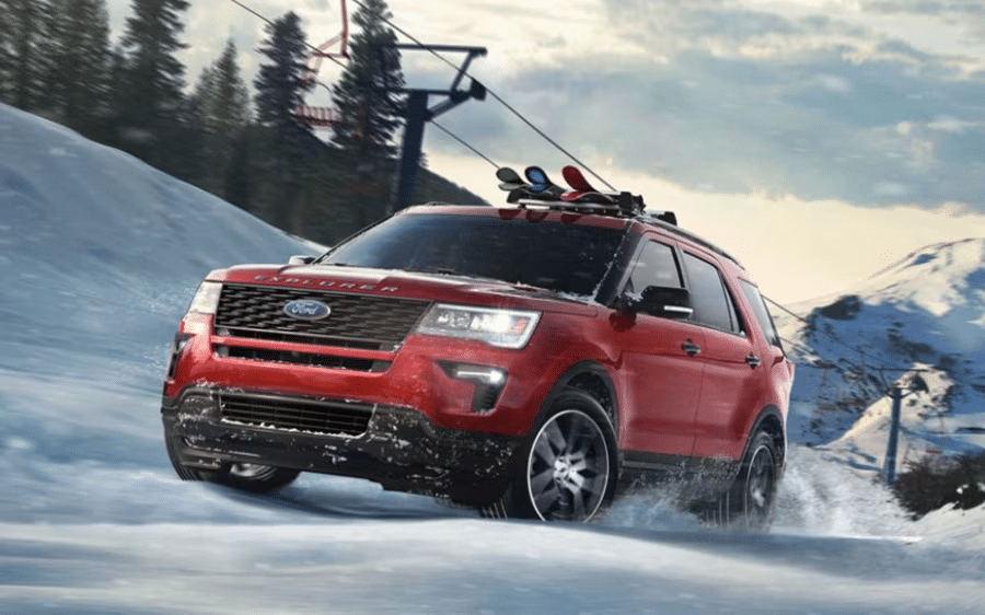 A red Ford Explorer SUV, one of the most popular Ford vehicles, drives up a snowy mountain road with snowboards mounted on its roof; pine trees and ski lift cables are visible in the background.