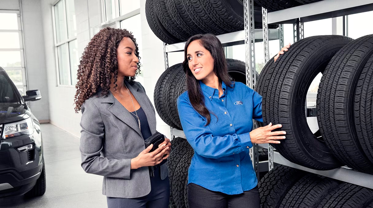 Two women stand by shelves of car tires in an auto shop. One, wearing a blue work shirt, demonstrates a tire to the other woman in business attire, reflecting excellent customer service. A black SUV and Ford vehicles are visible in the background.