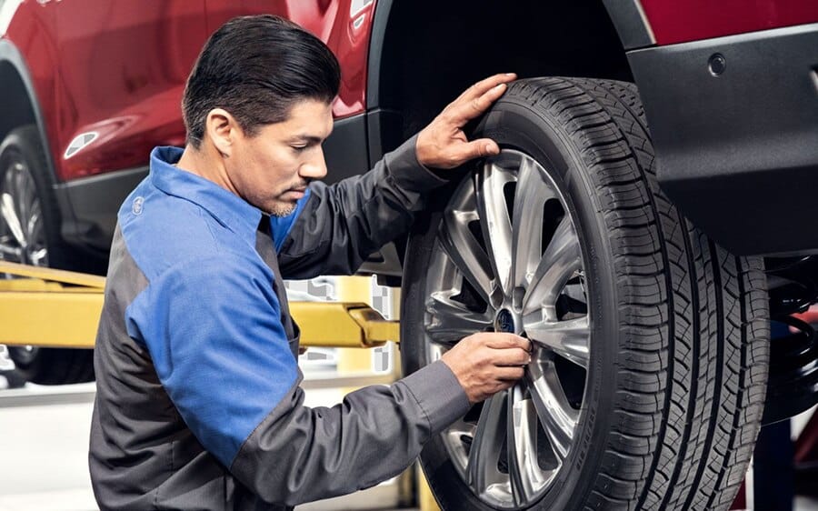 A mechanic stands beneath a red Ford truck lifted on a hydraulic car lift, inspecting or working on the vehicle’s underside with a strong focus on customer service.