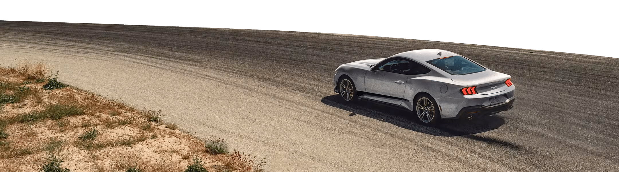 A silver sports car, reminiscent of high-performance Ford vehicles, drives on a curved asphalt racetrack bordered by dry grass and dirt.