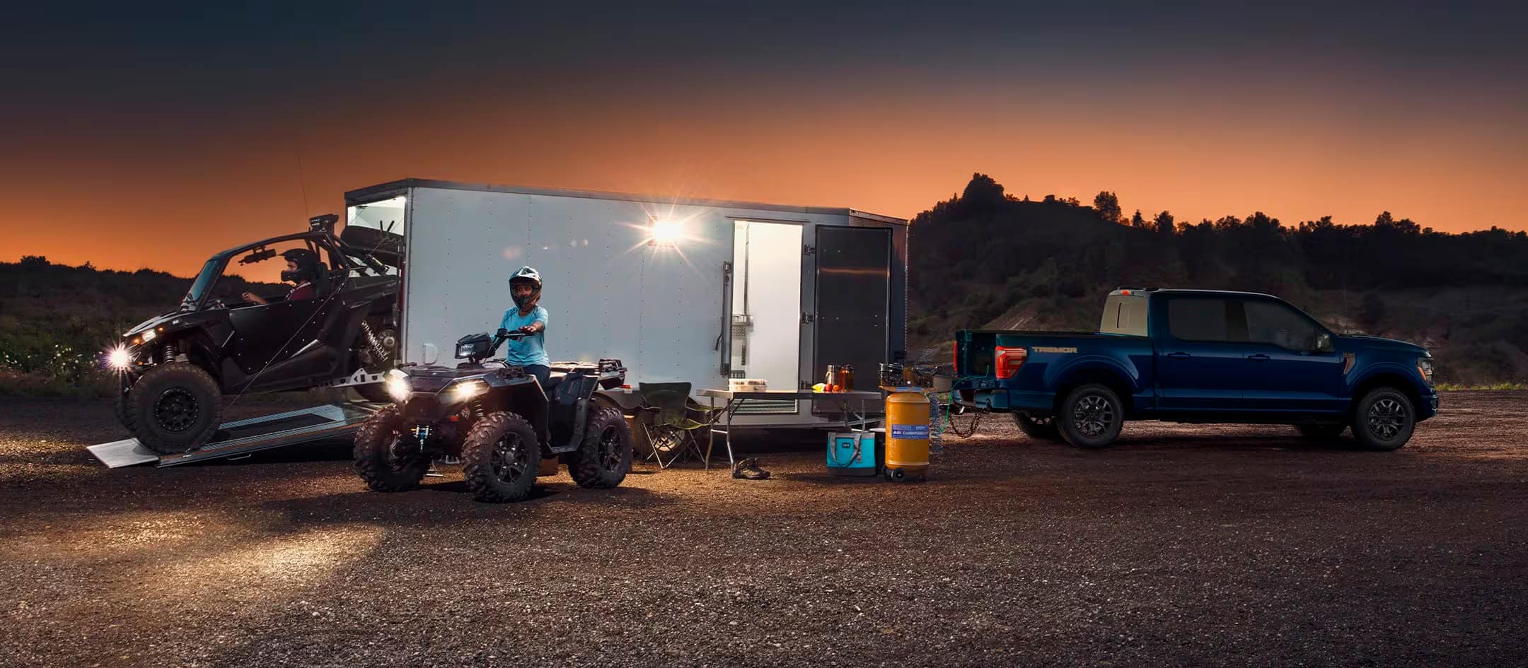 Two people drive ATVs from a trailer hitched to a parked F-150.