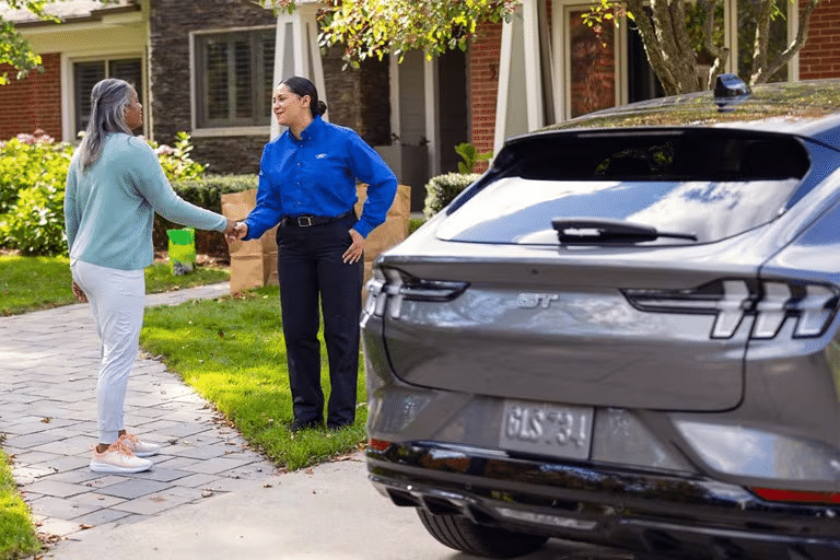 A Ford Pickup and Delivery representative is drops off a Ford Mustang Mach-E — greeting the owner in front of their home. 
