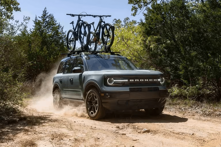 A Ford Bronco Sport SUV drives down a dirt road with two bikes mounted to a roof rack. 