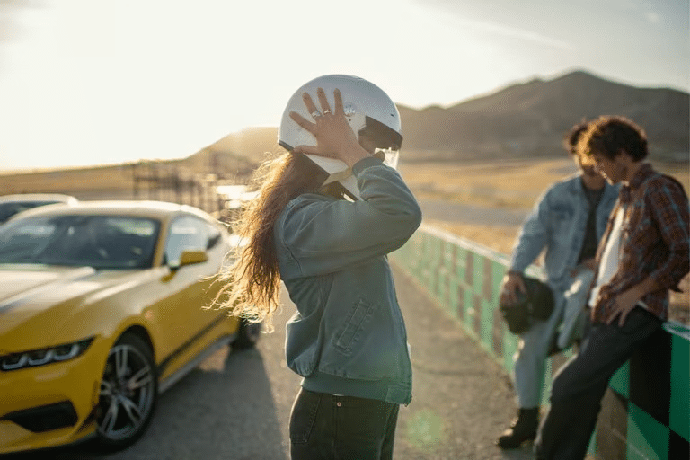 A woman puts on a racing helmet at a track. Two men and a Ford Mustang sit in the background. 