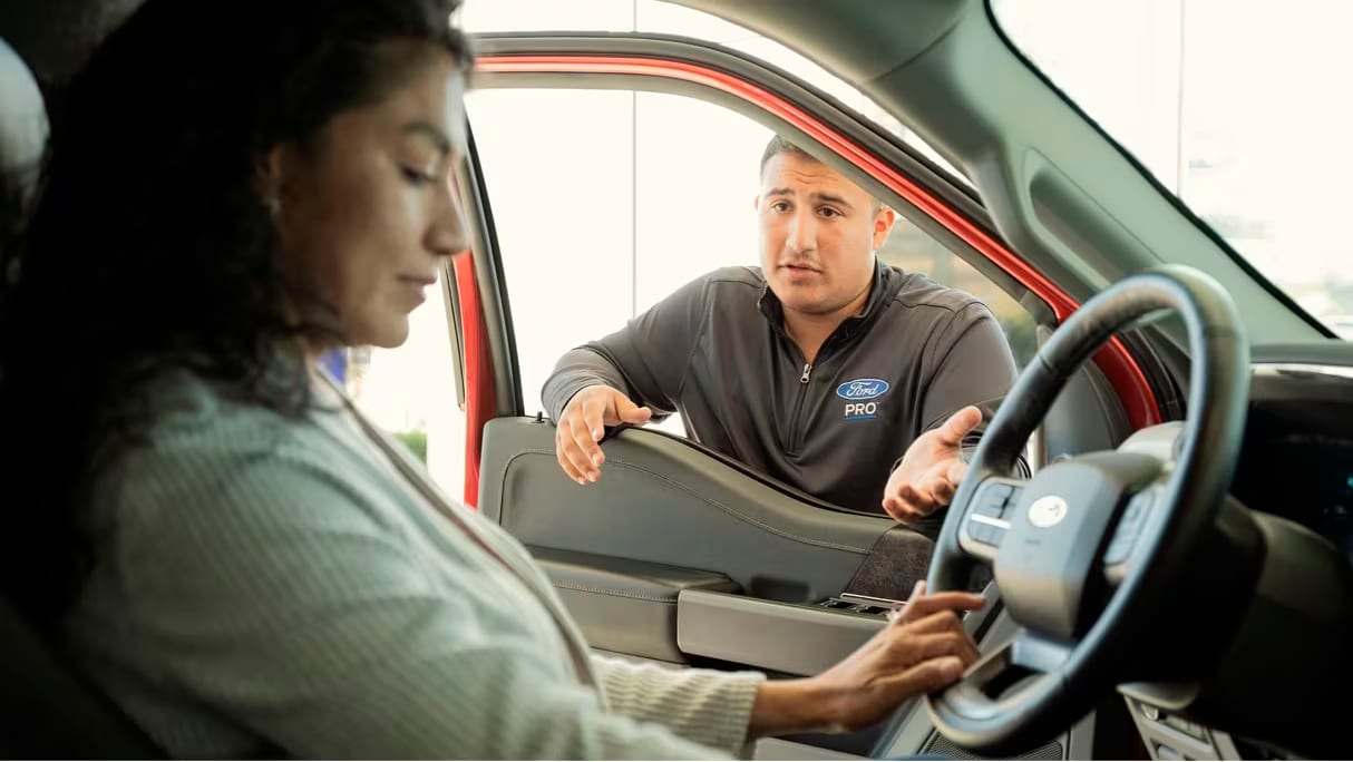In the foreground a woman is seated at the steering wheel of a Ford vehicle. A Ford Pro specialist is at the open window of a slightly ajar door