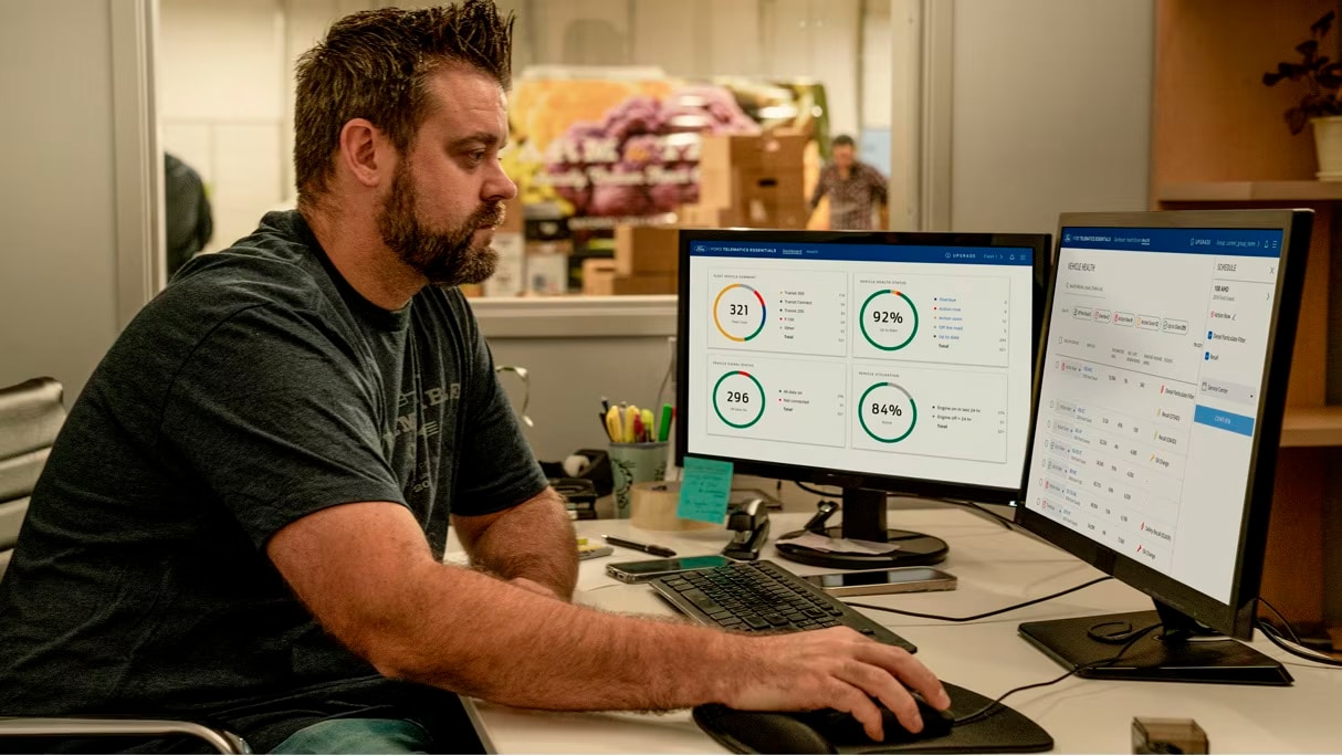 Man sitting at a desk in front of two computer monitors with his hand on the computer mouse. In the background there’s an employee, boxes and a van.