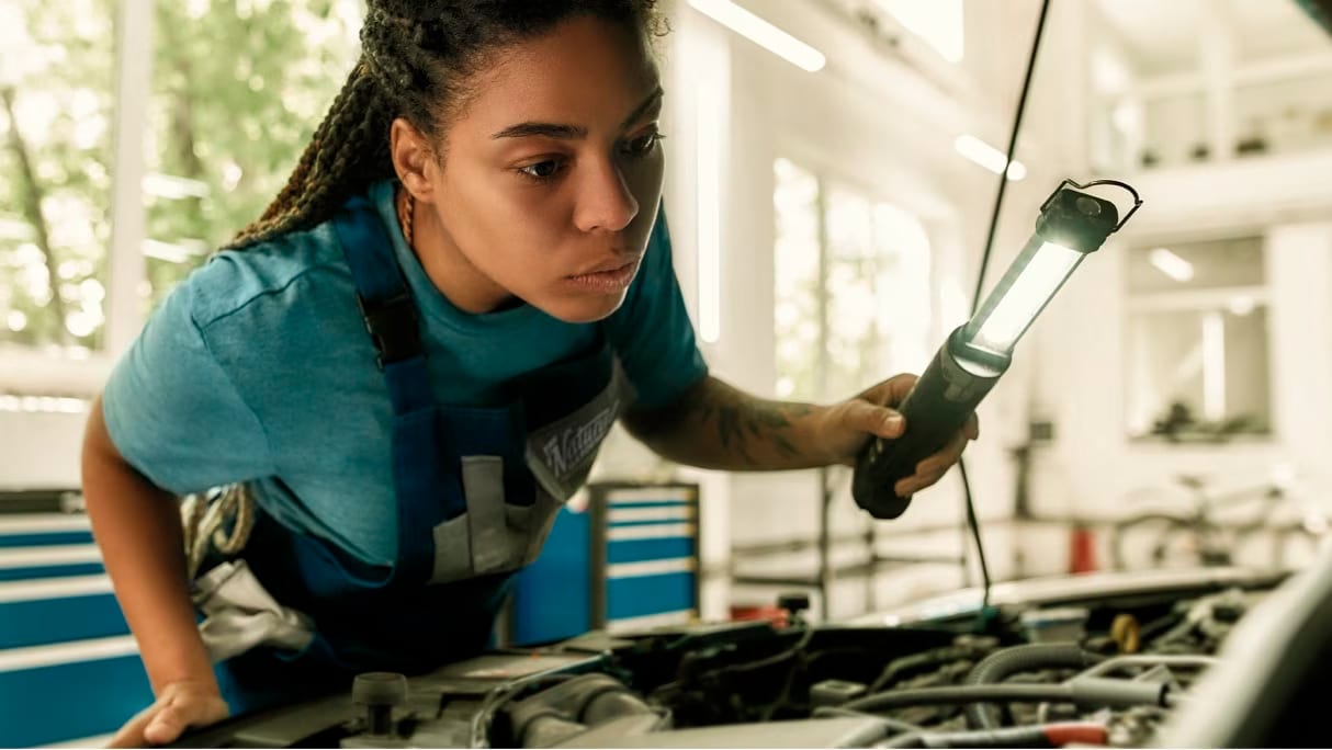 Ford Pro™ client in a garage looking under the hood of a vehicle. In her left hand she is holding a small lamp to illuminate the engine.