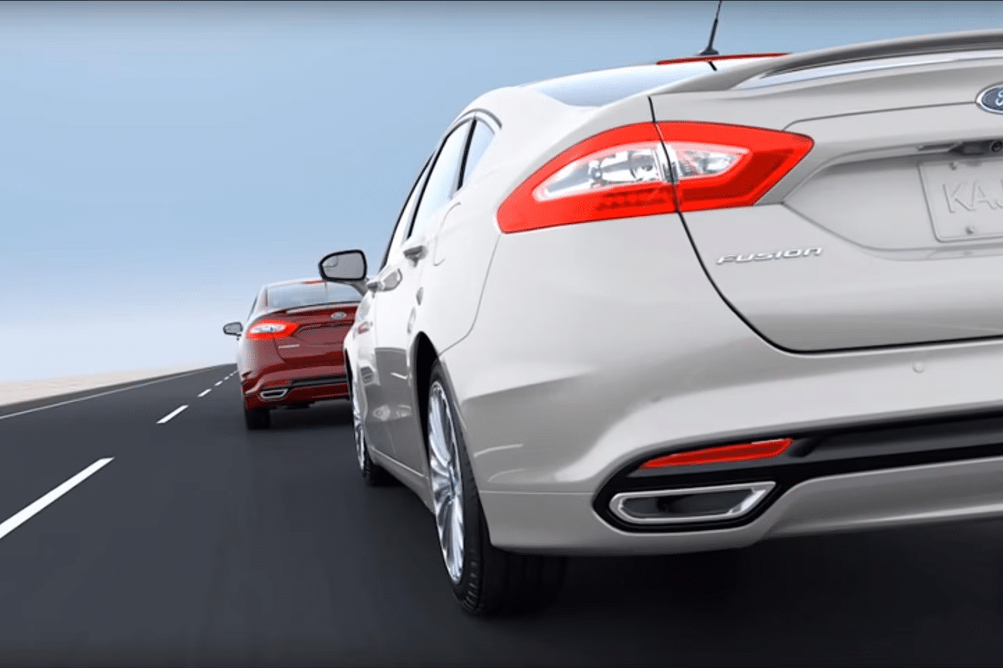 Two Ford Fusion cars, one white and one red, drive closely together on a multi-lane highway near Mississauga under a clear sky. The image is taken from behind at a low angle, focusing on the rear of the white Ford vehicle.