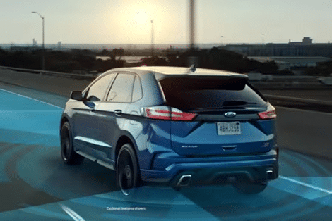 A blue SUV, part of the Ford vehicles lineup, drives on a highway at sunset as blue digital graphics illustrate advanced driver-assist or autonomous driving technology in use.
