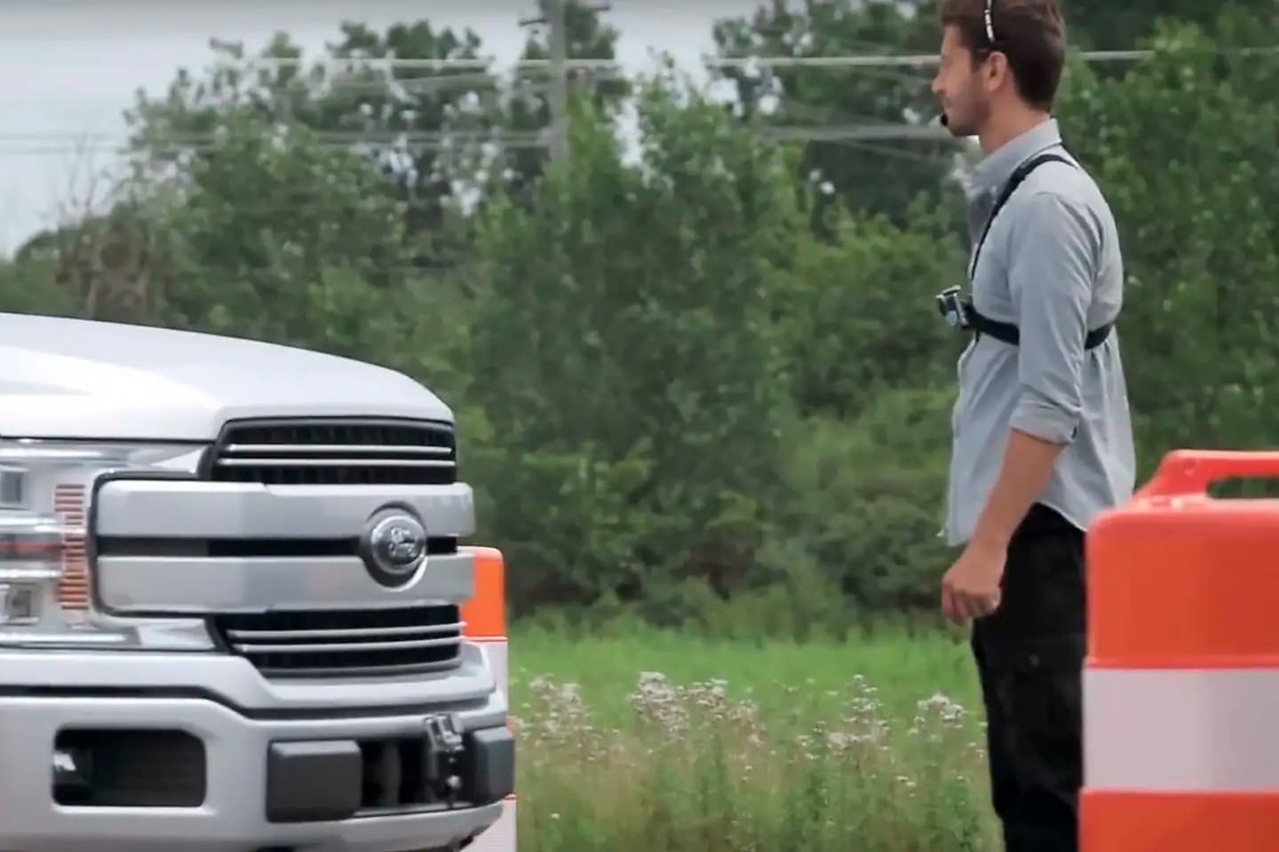 A man wearing a chest-mounted camera stands facing a white Ford pickup truck near orange-and-white barriers, exemplifying customer service in Mississauga. Green trees and grass provide a scenic background.